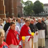 Uroczysta procesja z udziałem duchownych, w tym kanonika w czarnej sutannie, oraz wiernych zgromadzonych przed zabytkową budowlą.