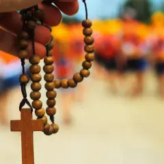 Drewniany różaniec w dłoni, symbol modlitwy za brata. W tle rozmyte sylwetki ludzi.