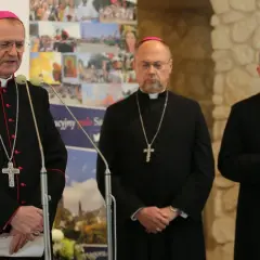 Trzech biskupów w czarnych szatach i z krzyżami na szyjach podczas zjazdu biskupów. Jeden z nich przemawia przy mikrofonie.