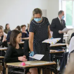 Nauczycielka w maseczce i rękawiczkach rozdaje arkusze egzaminacyjne uczniom siedzącym w ławkach.