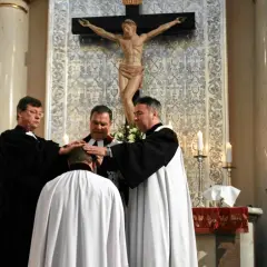 Trzech duchownych w szatach błogosławi klęczącego mężczyznę. Ceremonia w kościele protestanckim.