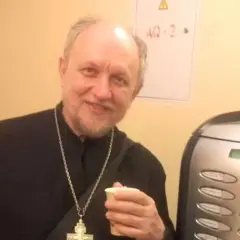 Uśmiechnięty священник w czarnej szacie z krzyżem na szyi trzyma kubek.