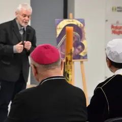 Muzułmański duchowny w białej czapce słucha przemówienia obok biskupa.