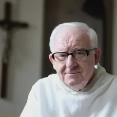 Starszy mężczyzna w okularach, w białej szacie, na tle krzyża. Może rozważa dogmat o nieomylności papieża.