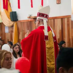 Biskup w czerwonej szacie i mitrze błogosławi wiernych. Jego laska biskupa jest ozdobiona złotymi krzyżami.
