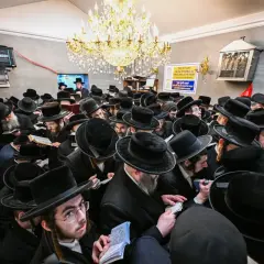 Tłum chasydów w czarnych kapeluszach i płaszczach, skupionych nad księgami, w sali ozdobionej żyrandolem.