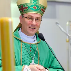 Uśmiechnięty prymas polski w zielonych szatach i mitrze, przemawia przy mikrofonie. W tle krzyż.
