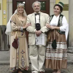 Ormianie w Polsce. Trzy osoby w strojach ludowych, dwie kobiety w tradycyjnych sukniach i mężczyzna w garniturze, pozują przed muzeum.