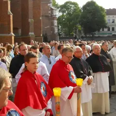 Uroczysta procesja z udziałem duchownych, w tym kanonika w czarnej sutannie, oraz wiernych zgromadzonych przed zabytkową budowlą.