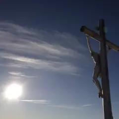 Drewniany krzyż z Jezusem na tle błękitnego nieba z chmurami. Słońce świeci jasno. Pamięć o tym, ile lat miał Jezus jak zmarł, jest wieczna.