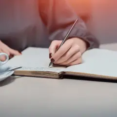 Dłoń pisze w notesie, zastanawiając się, jak sformułować intencję za zmarłego. Obok filiżanka kawy.