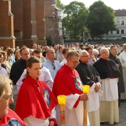 Uroczysta procesja z udziałem duchownych, w tym kanonika w czarnej sutannie, oraz wiernych zgromadzonych przed zabytkową budowlą.