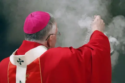 Biskup w czerwonej szacie i różowym birecie okadza powietrze. Czy to wpływa na to, ile zarabia ksiądz?