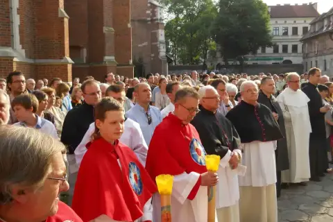 Uroczysta procesja z udziałem duchownych, w tym kanonika w czarnej sutannie, oraz wiernych zgromadzonych przed zabytkową budowlą.