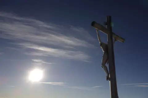 Drewniany krzyż z Jezusem na tle błękitnego nieba z chmurami. Słońce świeci jasno. Pamięć o tym, ile lat miał Jezus jak zmarł, jest wieczna.