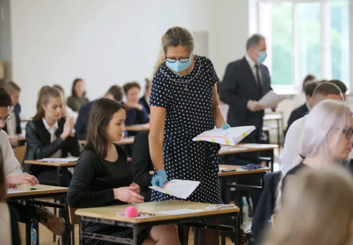 Nauczycielka w maseczce i rękawiczkach rozdaje arkusze egzaminacyjne uczniom siedzącym w ławkach.
