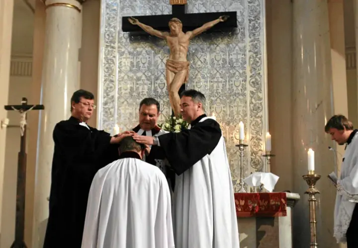 Trzech duchownych w szatach błogosławi klęczącego mężczyznę. Ceremonia w kościele protestanckim.