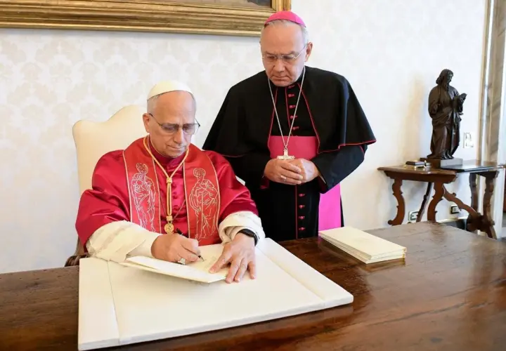 Biskup w purpurowej szacie podpisuje dokument, podczas gdy inny duchowny w czerni i różu stoi obok, obserwując. To ważny moment, być może podpisanie adhortacji.