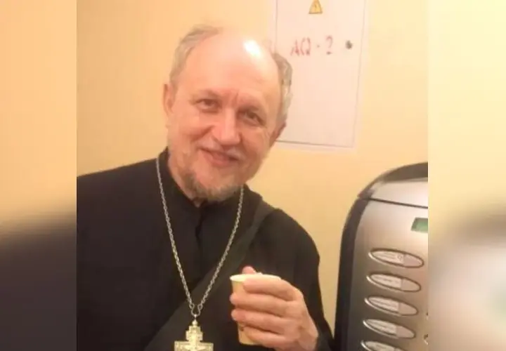 Uśmiechnięty священник w czarnej szacie z krzyżem na szyi trzyma kubek.
