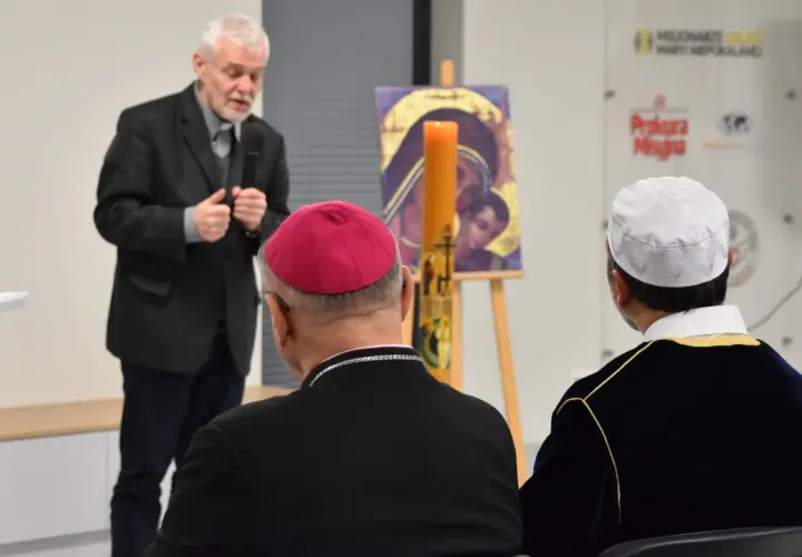 Muzułmański duchowny w białej czapce słucha przemówienia obok biskupa.