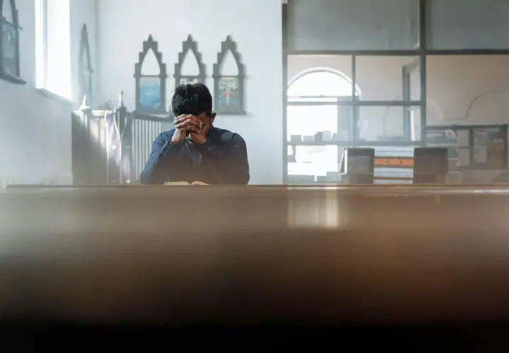 Mężczyzna w kościele pogrążony w głębokiej kontemplacji, z rękami złożonymi na twarzy.