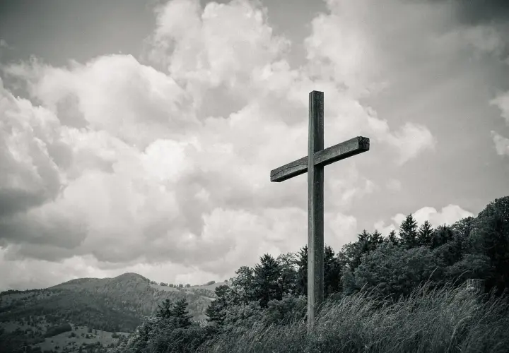Drewniany krzyż na tle burzowych chmur, niczym biblijna góra, góruje nad zalesionym zboczem.