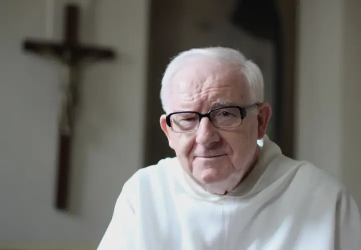 Starszy mężczyzna w okularach, w białej szacie, na tle krzyża. Może rozważa dogmat o nieomylności papieża.