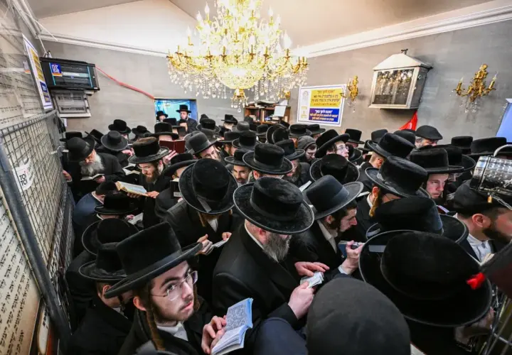 Tłum chasydów w czarnych kapeluszach i płaszczach, skupionych nad księgami, w sali ozdobionej żyrandolem.