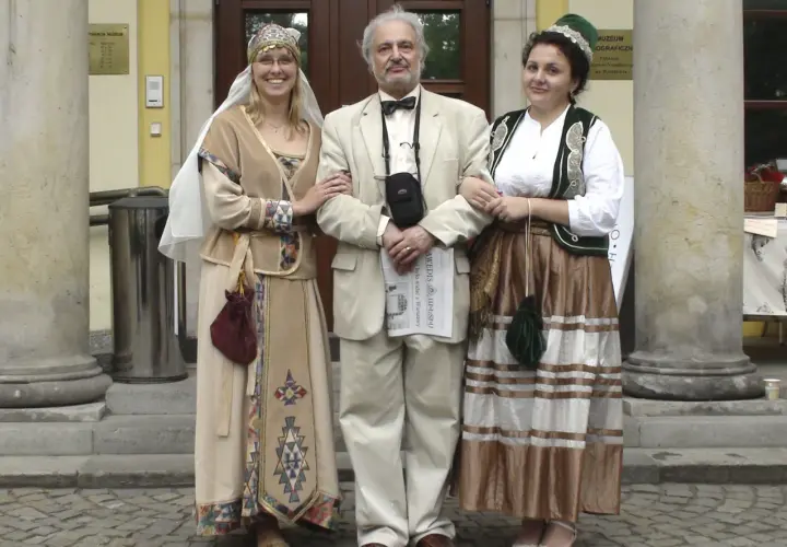 Ormianie w Polsce. Trzy osoby w strojach ludowych, dwie kobiety w tradycyjnych sukniach i mężczyzna w garniturze, pozują przed muzeum.