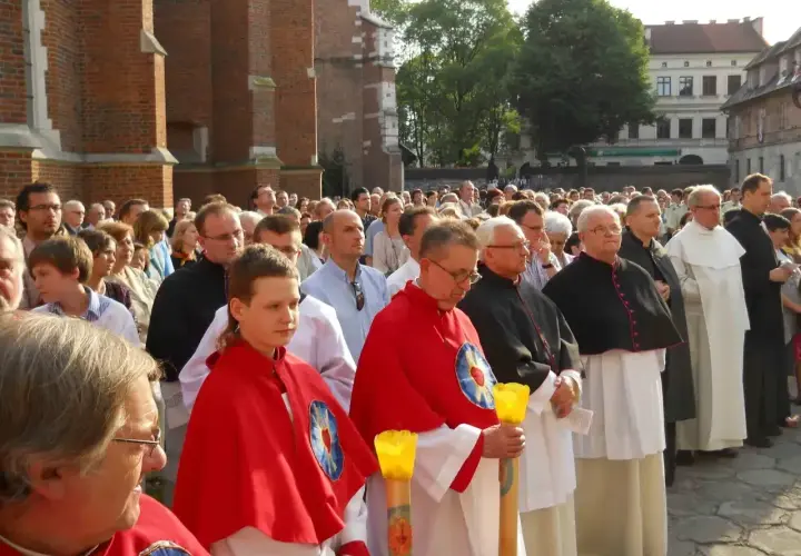Uroczysta procesja z udziałem duchownych, w tym kanonika w czarnej sutannie, oraz wiernych zgromadzonych przed zabytkową budowlą.