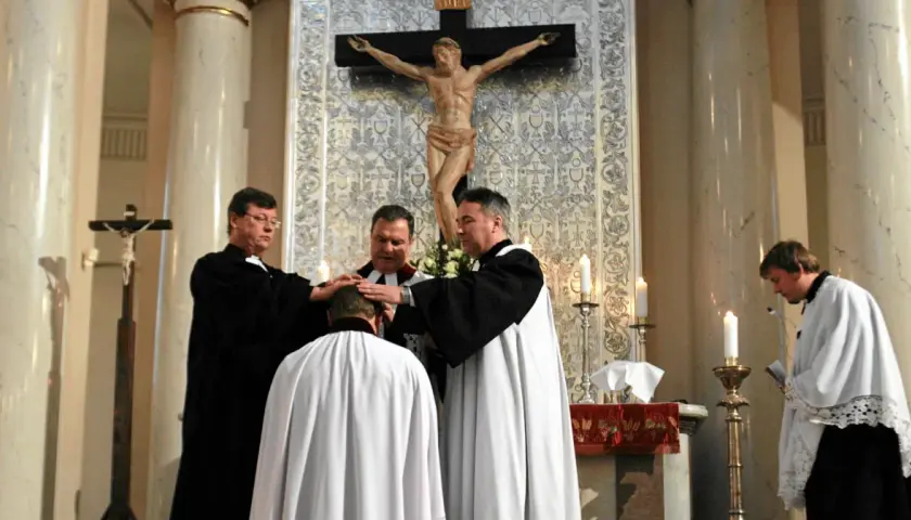 Trzech duchownych w szatach błogosławi klęczącego mężczyznę. Ceremonia w kościele protestanckim.