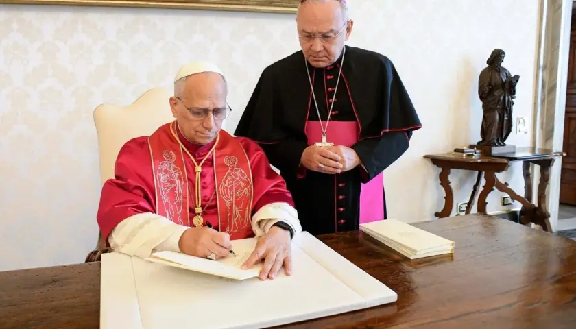 Biskup w purpurowej szacie podpisuje dokument, podczas gdy inny duchowny w czerni i różu stoi obok, obserwując. To ważny moment, być może podpisanie adhortacji.