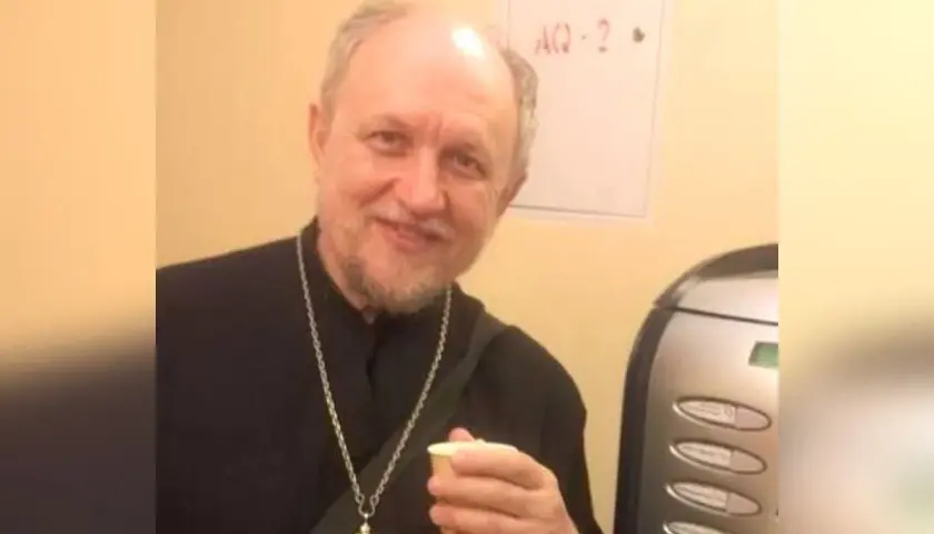 Uśmiechnięty священник w czarnej szacie z krzyżem na szyi trzyma kubek.