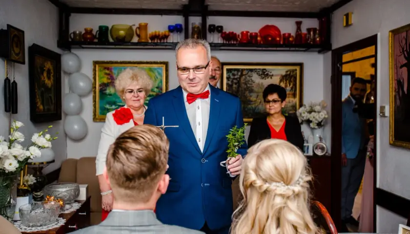 Uroczysty błogosławieństwo tekst. Mężczyzna w niebieskiej marynarce z czerwoną muszką trzyma krzyż i gałązkę.