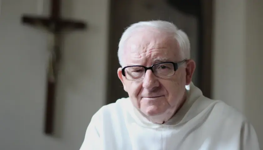 Starszy mężczyzna w okularach, w białej szacie, na tle krzyża. Może rozważa dogmat o nieomylności papieża.