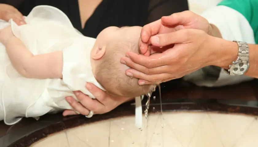 Chrzest dziecka w białej szacie. Czy ksiądz może odmówić chrztu nieślubnemu dziecku? Woda spływa z rąk do chrzcielnicy.