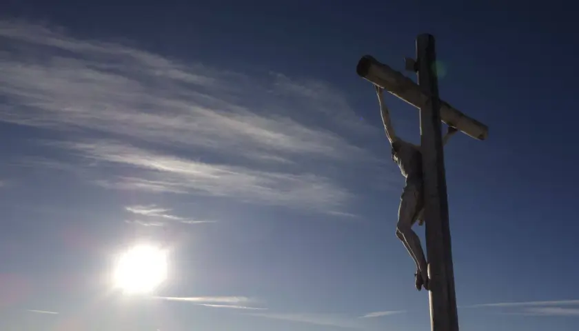 Drewniany krzyż z Jezusem na tle błękitnego nieba z chmurami. Słońce świeci jasno. Pamięć o tym, ile lat miał Jezus jak zmarł, jest wieczna.