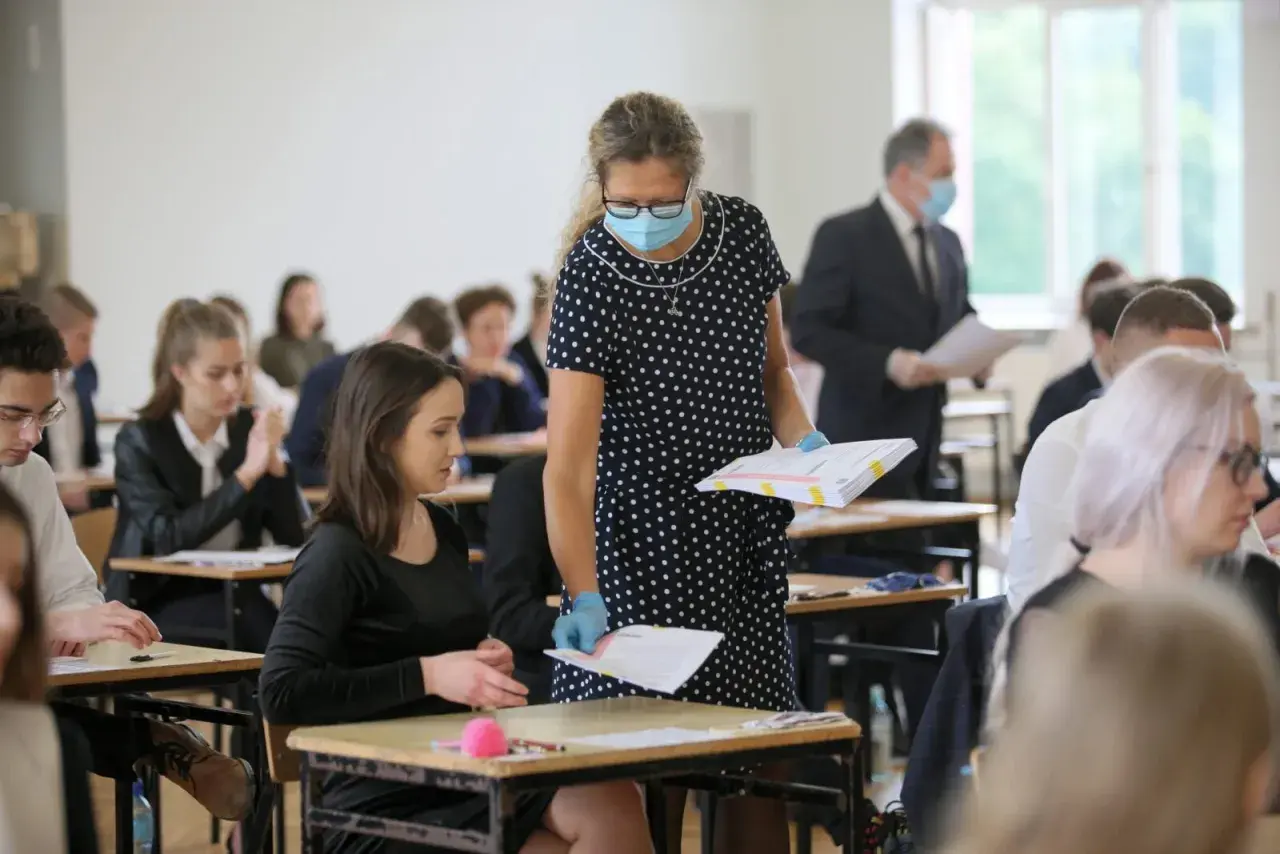 Nauczycielka w maseczce i rękawiczkach rozdaje arkusze egzaminacyjne uczniom siedzącym w ławkach.