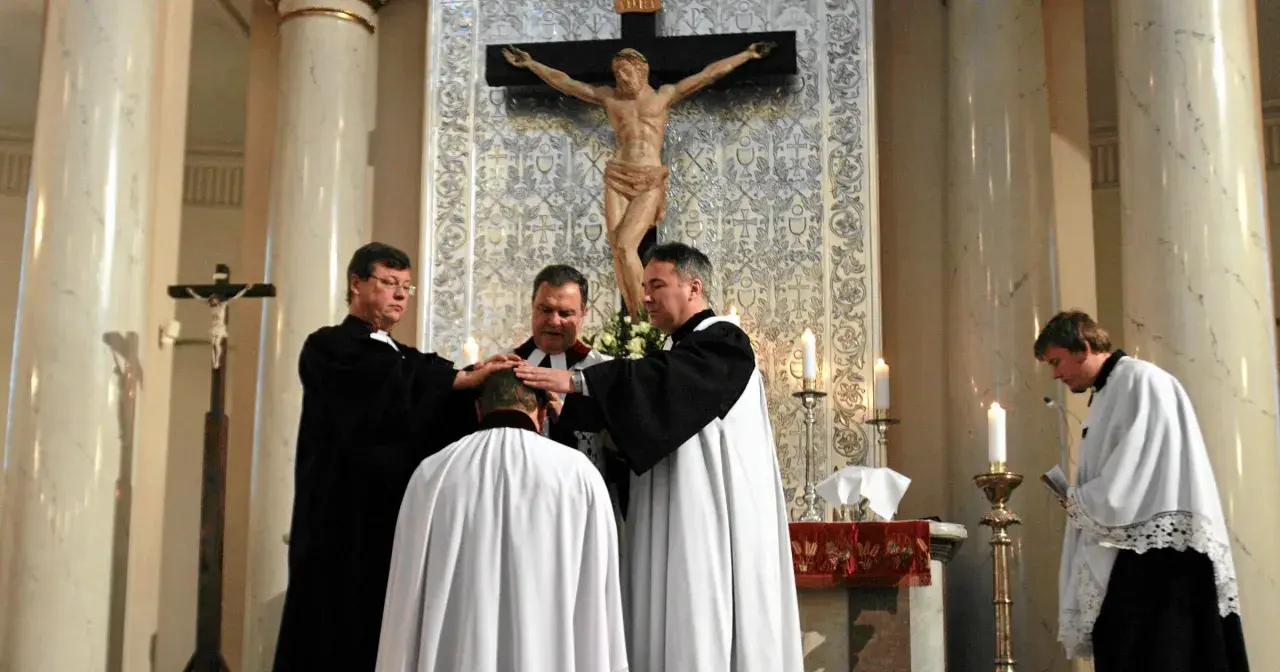 Trzech duchownych w szatach błogosławi klęczącego mężczyznę. Ceremonia w kościele protestanckim.