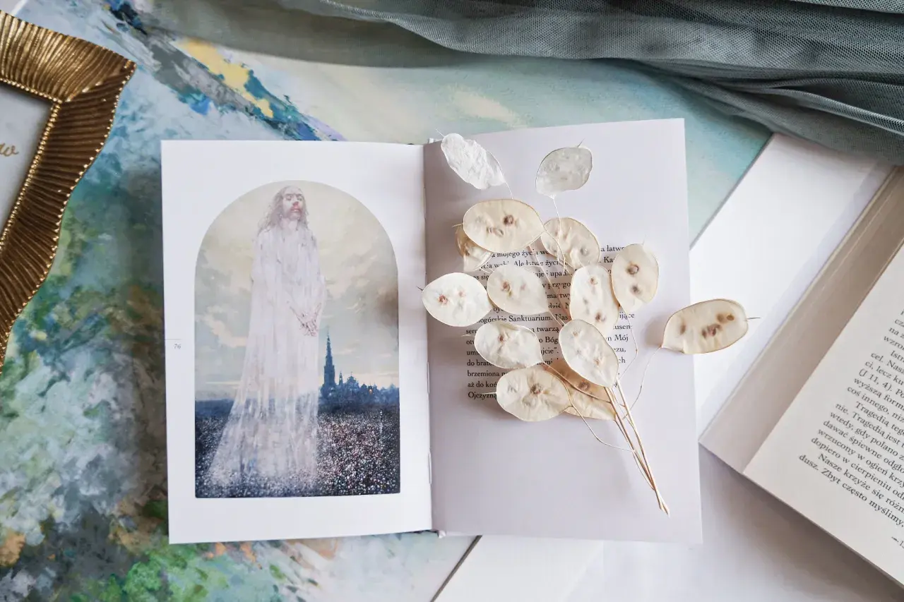 Otwarta księga z ilustracją postaci w bieli i suszonymi lunariami. Obok tekst, który może zawierać najpiękniejsze rozważania drogi krzyżowej.