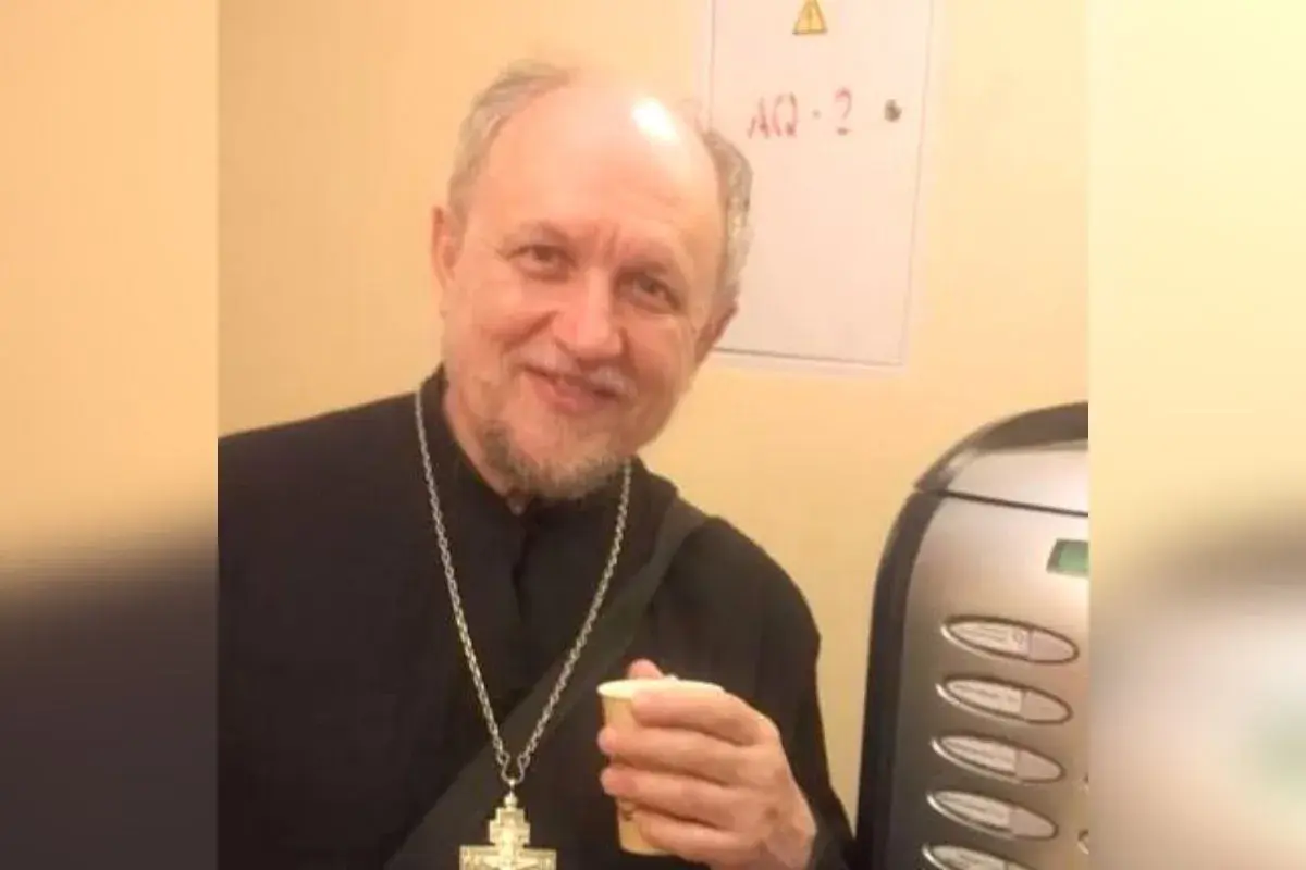 Uśmiechnięty священник w czarnej szacie z krzyżem na szyi trzyma kubek.