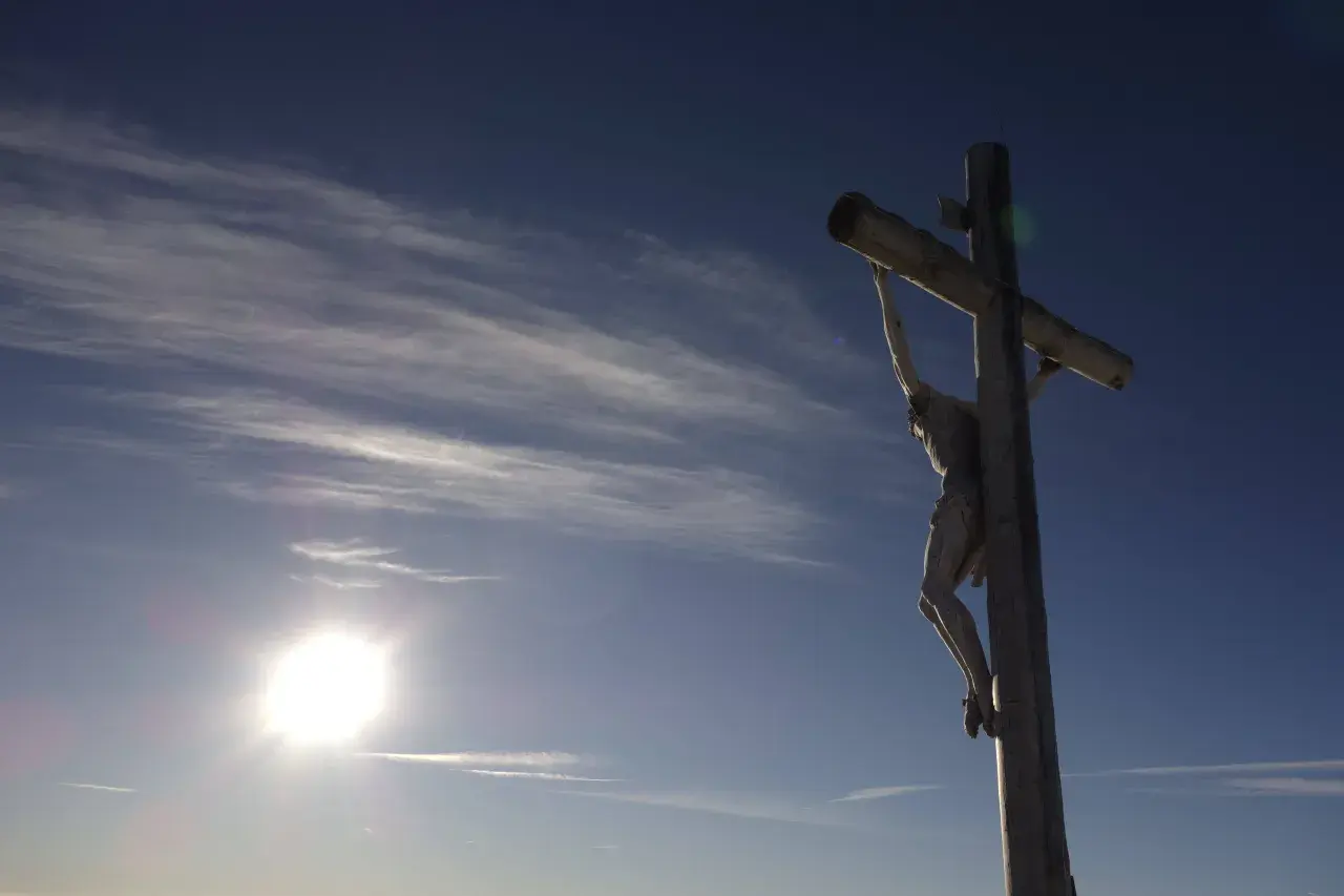 Drewniany krzyż z Jezusem na tle błękitnego nieba z chmurami. Słońce świeci jasno. Pamięć o tym, ile lat miał Jezus jak zmarł, jest wieczna.