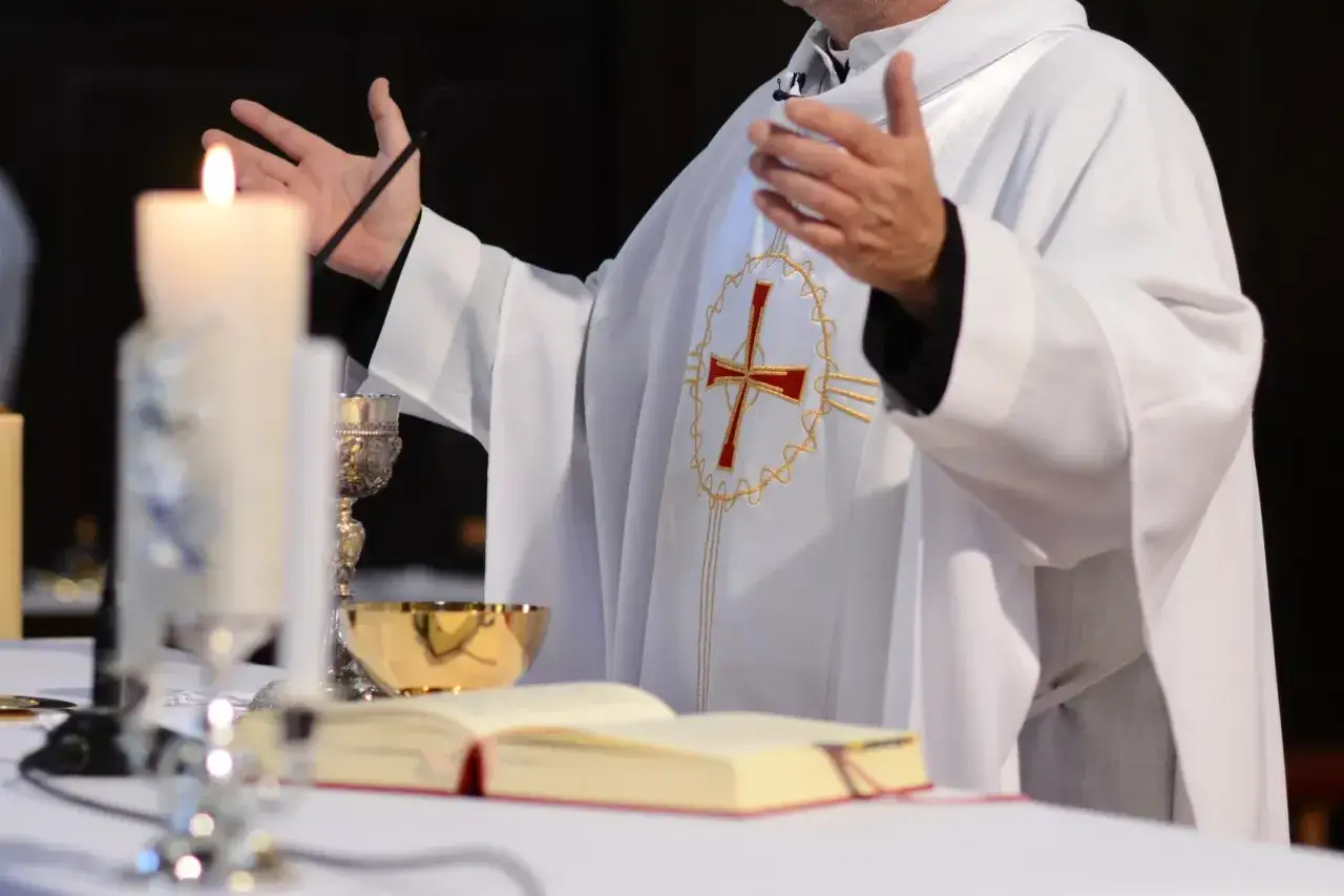 Ksiądz w białej albie z krzyżem, z rękami uniesionymi w geście modlitwy. Obok świeca i kielich. Czy ksiądz może mieć żonę? To pytanie nurtuje wielu.