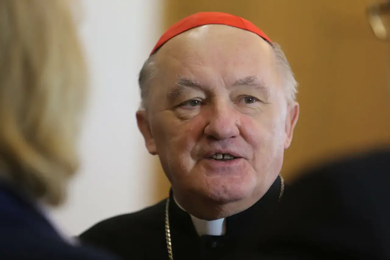 Uśmiechnięty biskup Nycz w czerwonej zuce i czarnej sutannie, w otoczeniu innych osób.