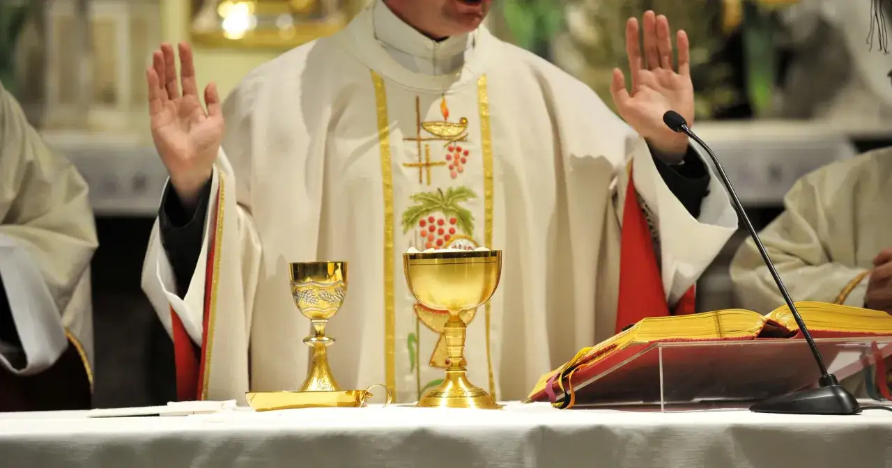 Kapłan w uroczystej szacie, z podniesionymi rękami, podczas mszy. Na ołtarzu kielichy i księga, symbol wyznania wiary.