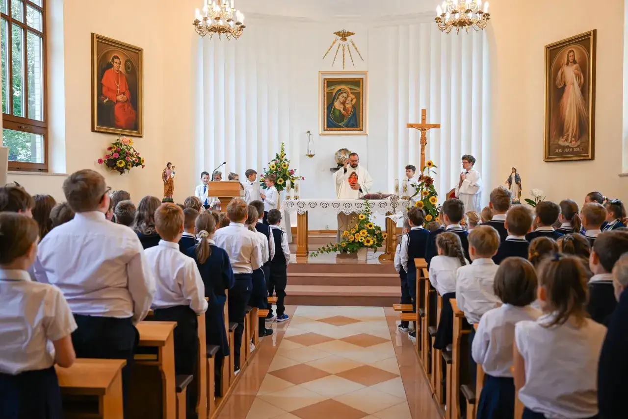 Duchowny w białej szacie odprawia mszę dla dzieci w kościele. Dzieci w strojach komunijnych stoją w ławkach.