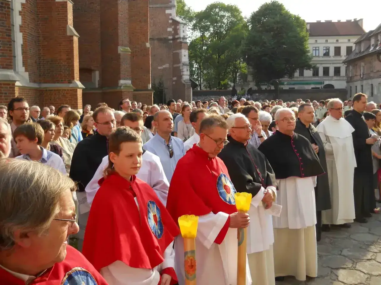 Uroczysta procesja z udziałem duchownych, w tym kanonika w czarnej pelerynie, oraz wiernych zgromadzonych przed zabytkową ceglaną budowlą.